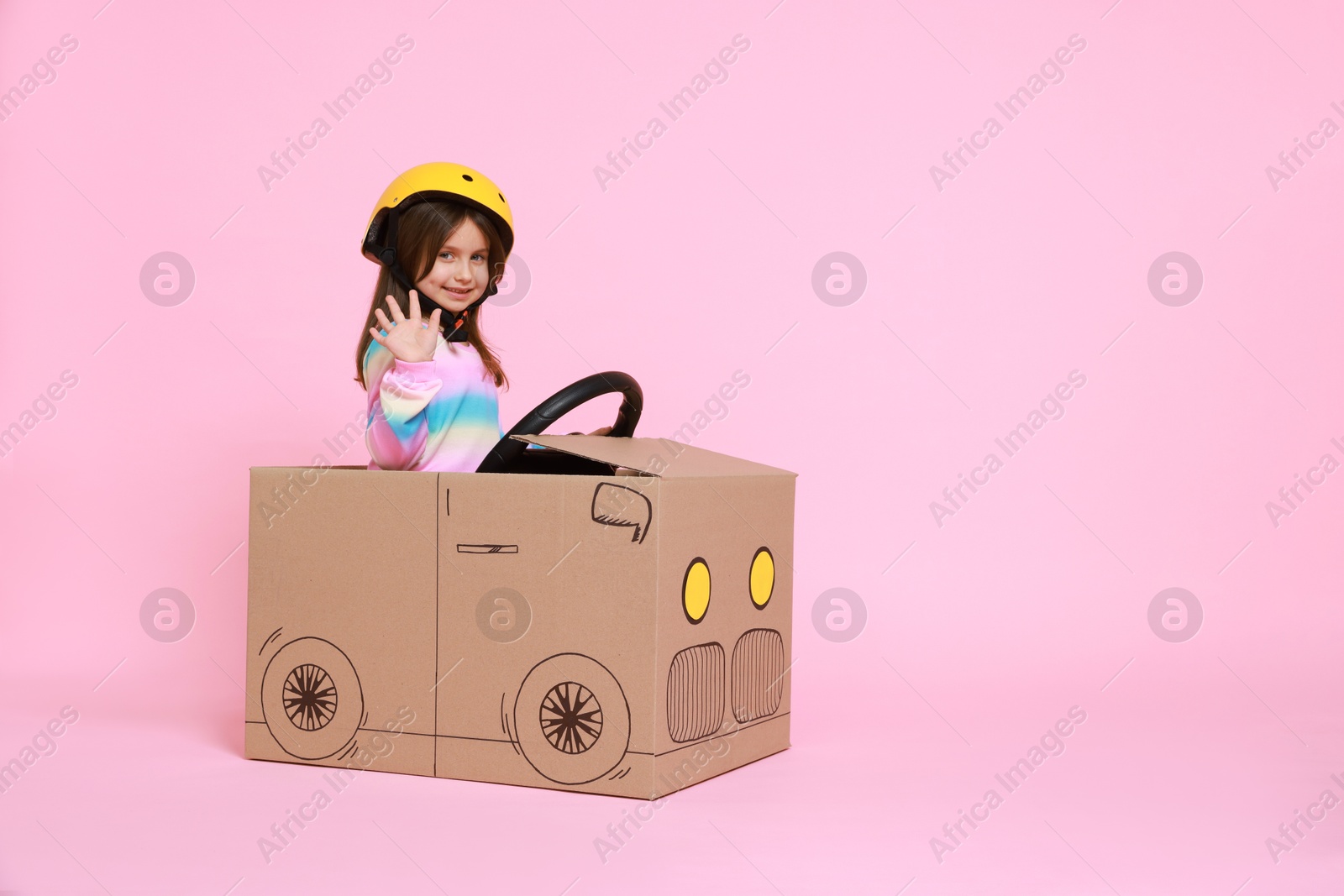 Photo of Little girl waving while driving car made with cardboard on light pink background