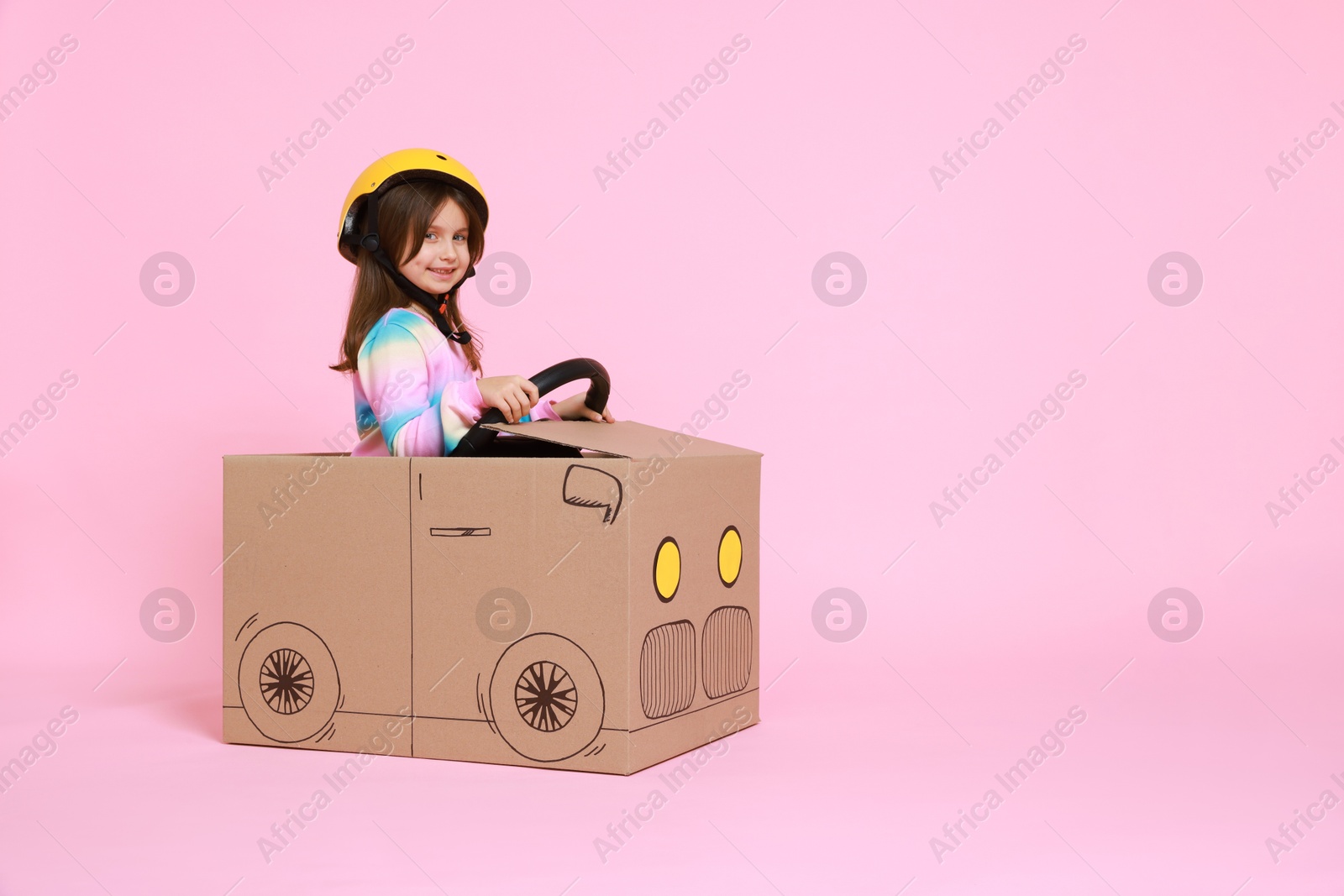 Photo of Little girl driving car made with cardboard on light pink background. Space for text