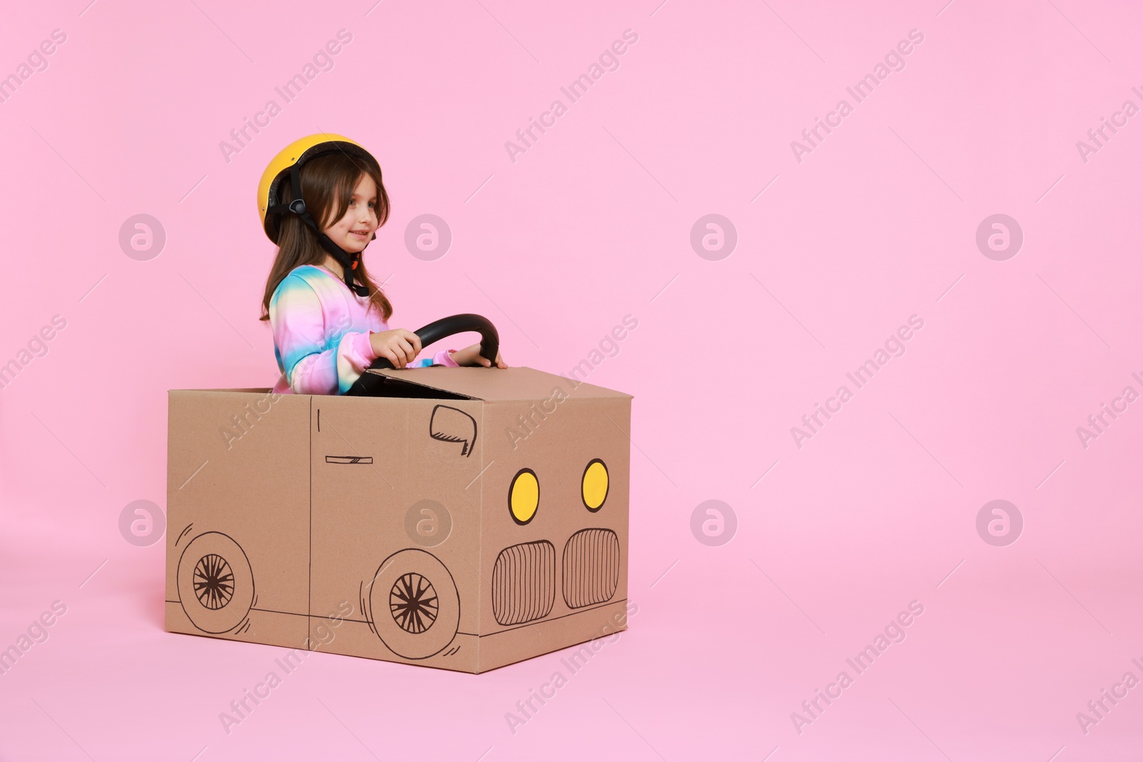 Photo of Little girl driving car made with cardboard on light pink background. Space for text