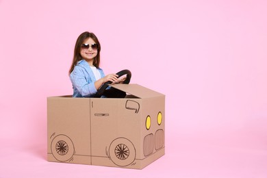 Little girl in sunglasses driving car made with cardboard on light pink background. Space for text