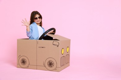 Little girl waving while driving car made with cardboard on light pink background. Space for text