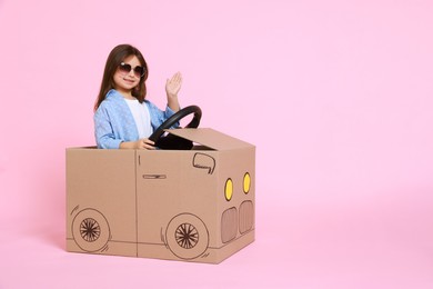 Little girl waving while driving car made with cardboard on light pink background. Space for text