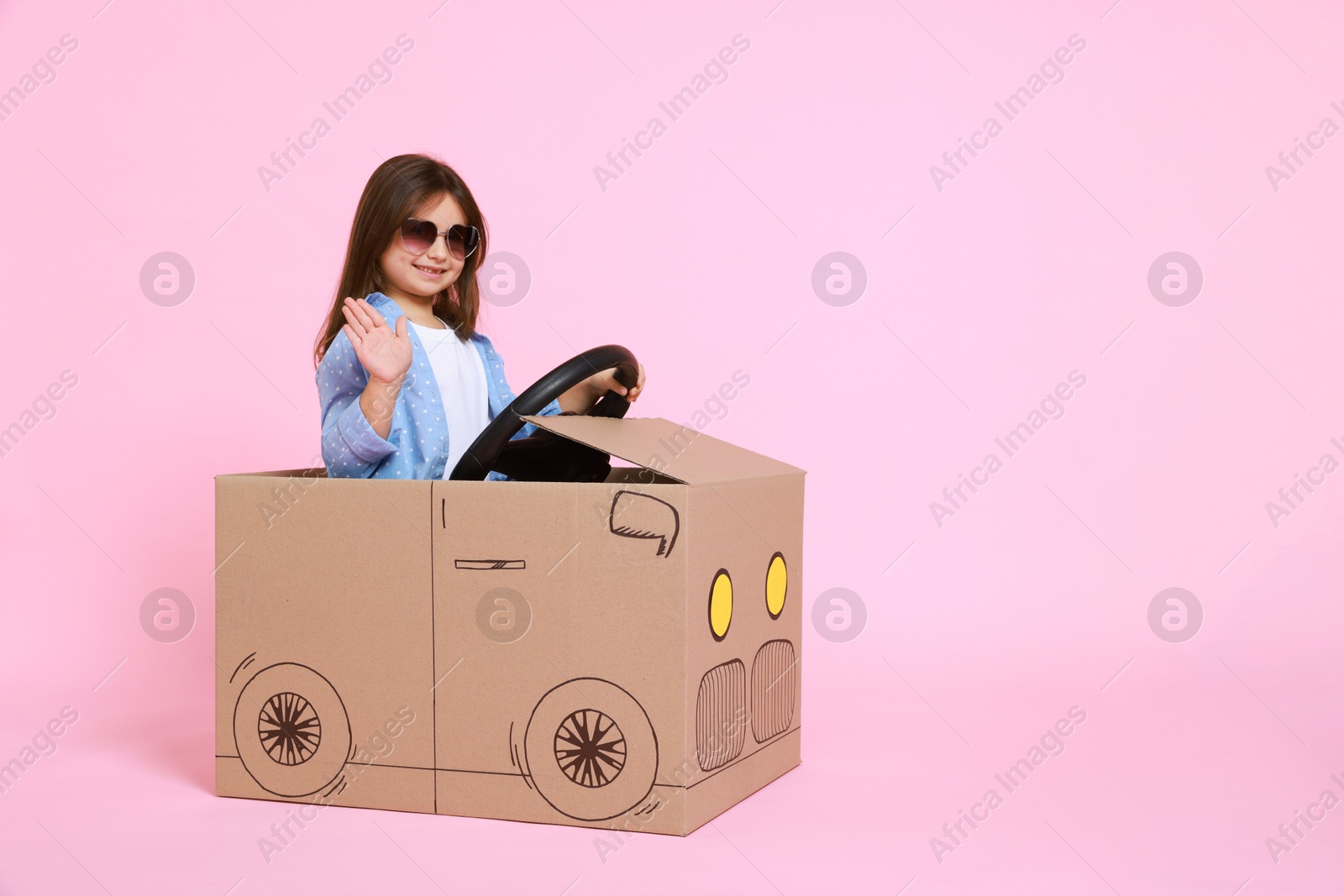 Photo of Little girl waving while driving car made with cardboard on light pink background. Space for text