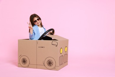 Little girl showing V-sign while driving car made with cardboard on light pink background. Space for text