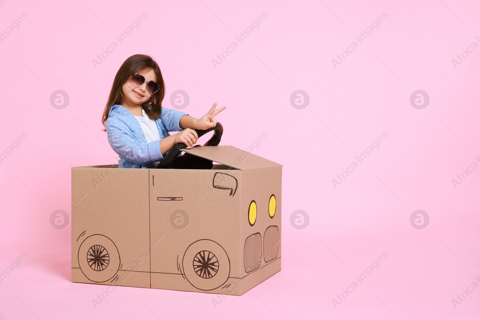 Photo of Little girl showing V-sign while driving car made with cardboard on light pink background. Space for text