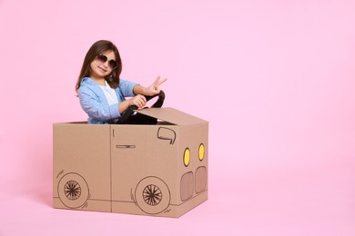 Little girl showing V-sign while driving car made with cardboard on light pink background. Space for text