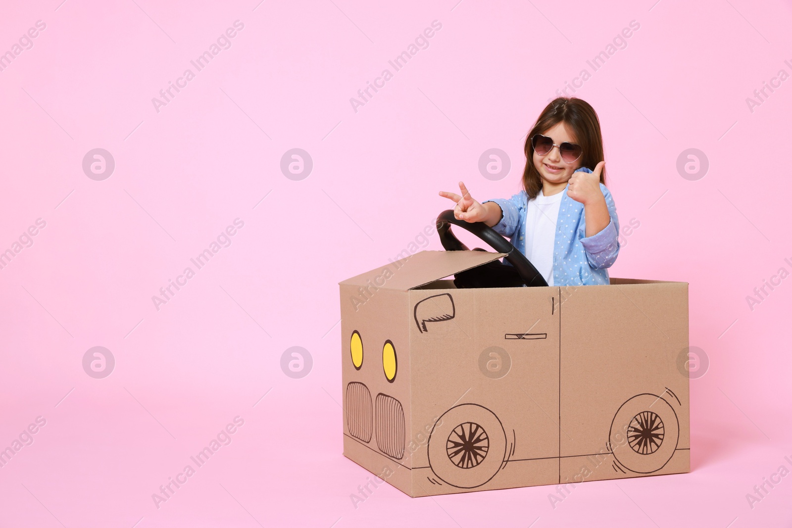 Photo of Little girl showing thumbs up while driving car made with cardboard on light pink background. Space for text