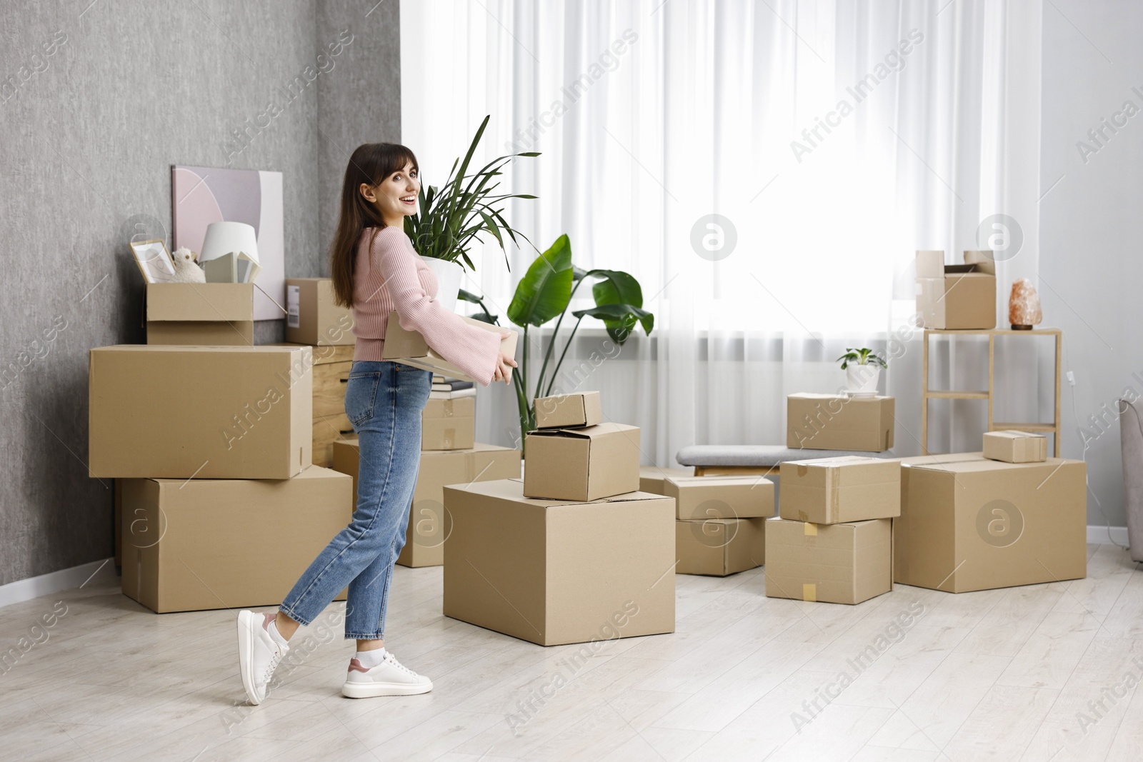 Photo of Moving day. Happy woman with cardboard box and houseplant in her new home