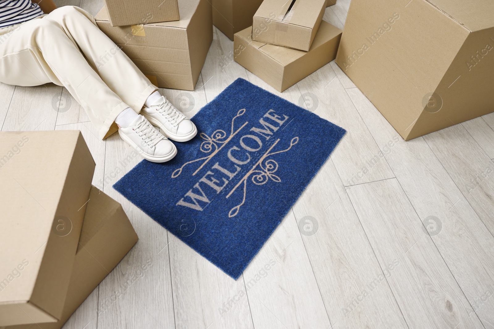 Photo of Moving day. Woman sitting near cardboard boxes and doormat with word Welcome on floor in her new home, closeup