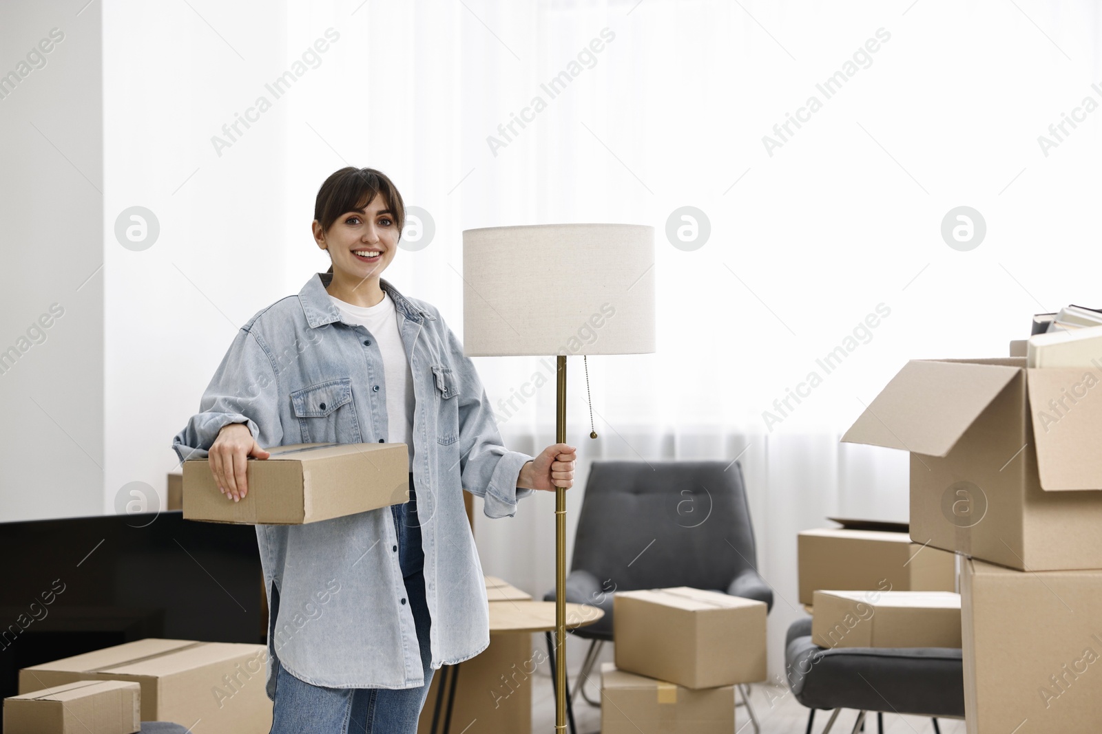 Photo of Moving day. Happy woman with lamp and cardboard box in her new home