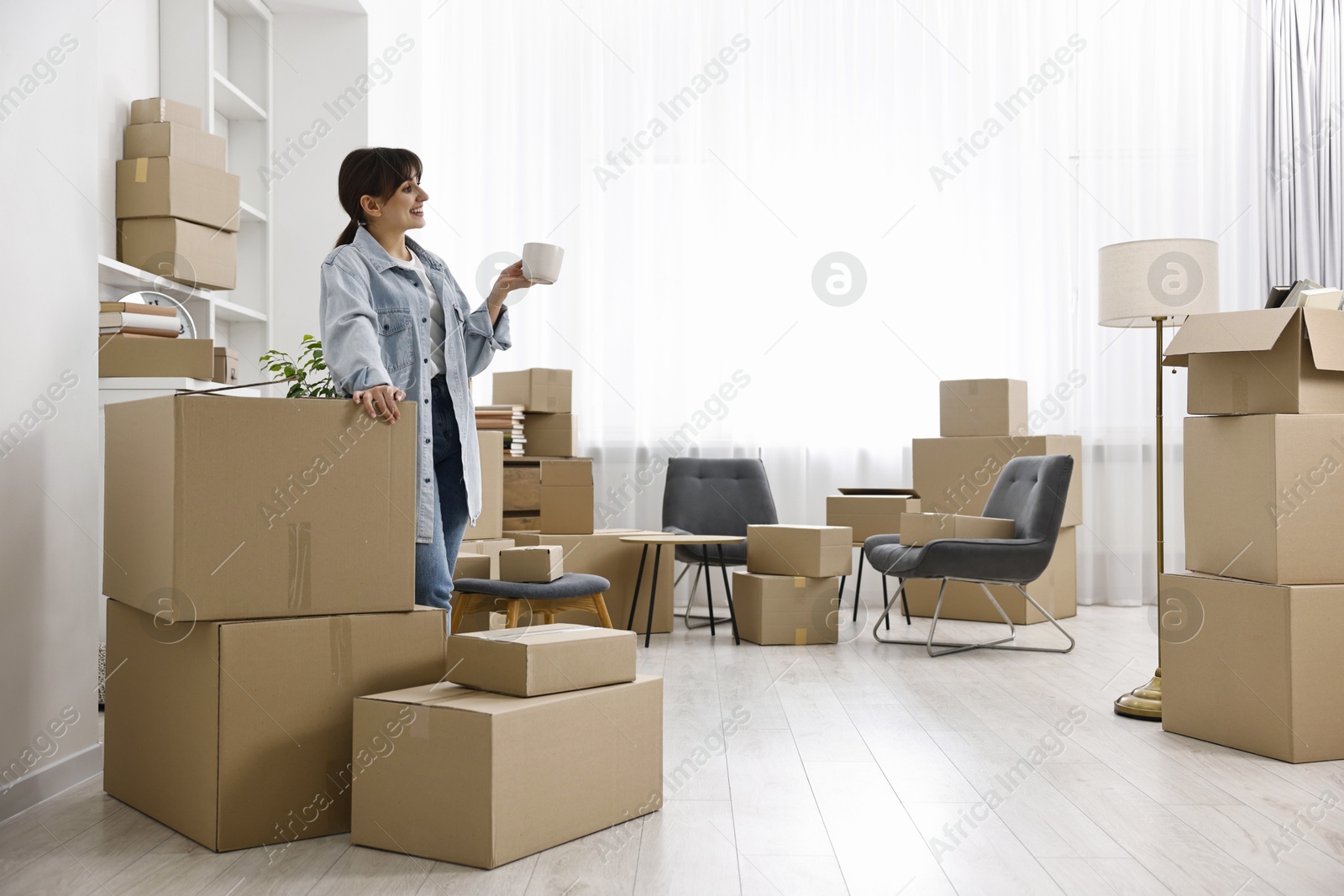 Photo of Moving day. Happy woman with cup of drink and cardboard boxes in her new home