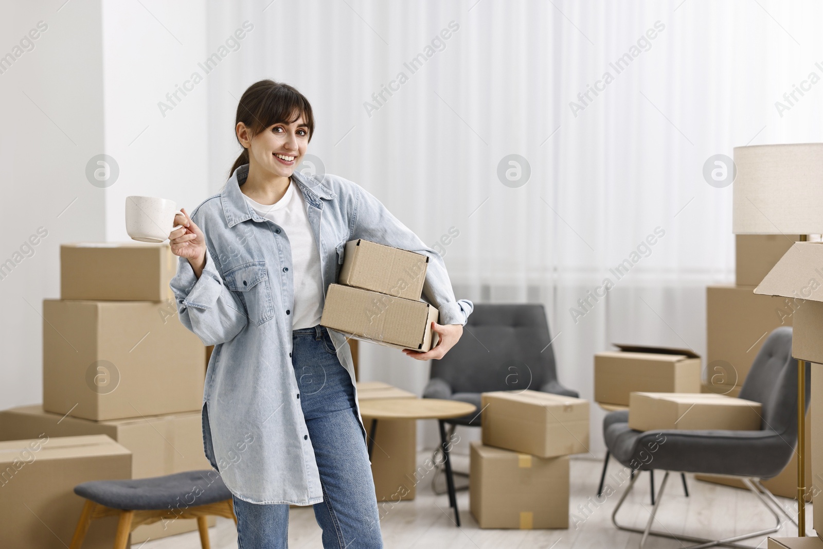 Photo of Moving day. Happy woman with cup of drink and cardboard boxes in her new home, space for text