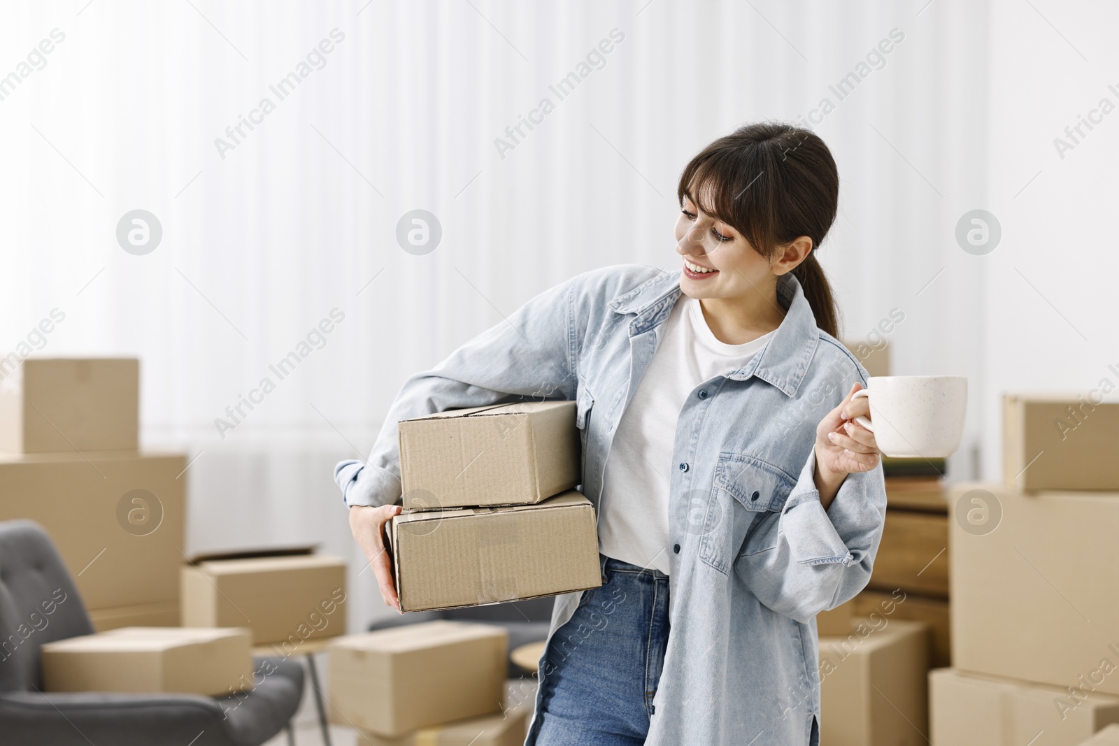 Photo of Moving day. Happy woman with cup of drink and cardboard boxes in her new home, space for text