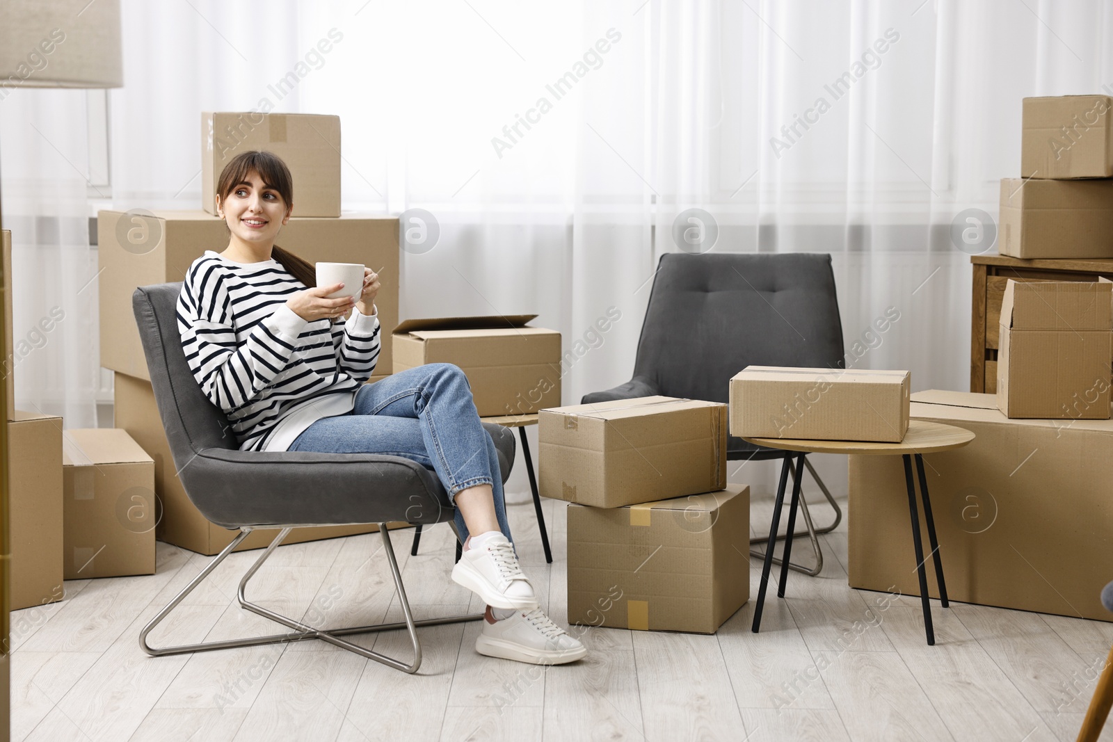 Photo of Moving day. Happy woman with cup of drink and cardboard boxes in new home