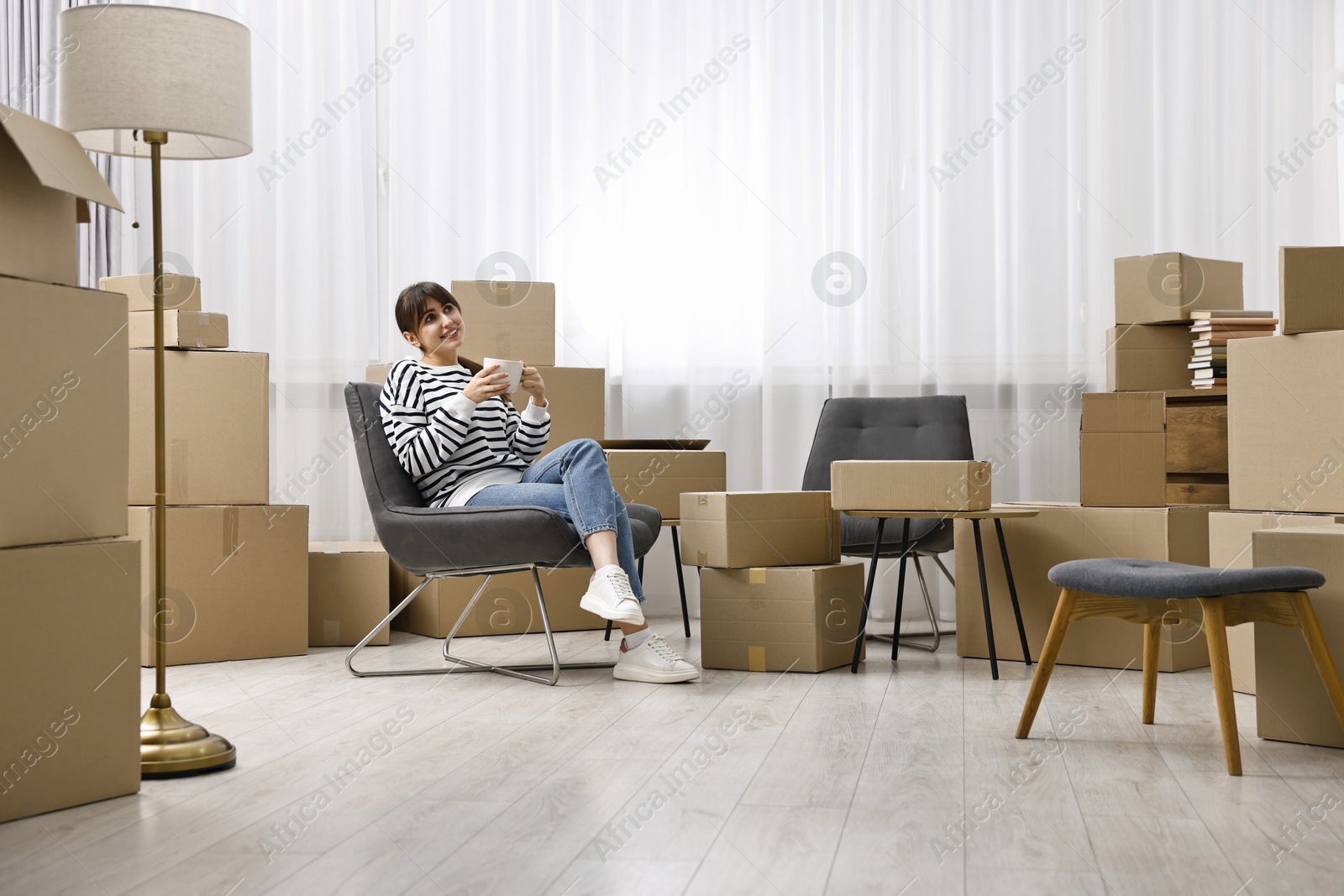 Photo of Moving day. Happy woman with cup of drink and cardboard boxes in new home