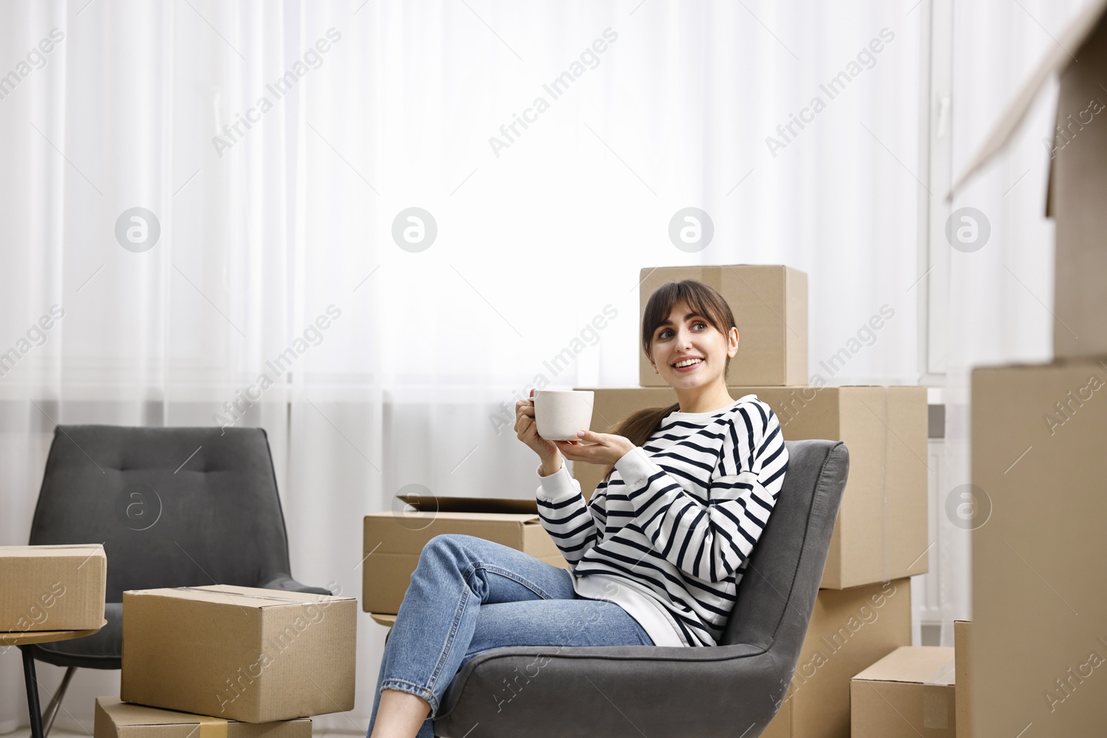 Photo of Moving day. Happy woman with cup of drink and cardboard boxes in new home