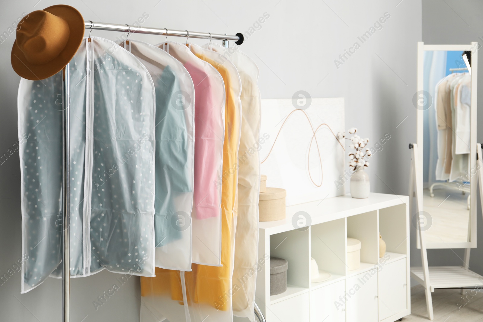 Photo of Garment covers with clothes and hat on rack near grey wall indoors