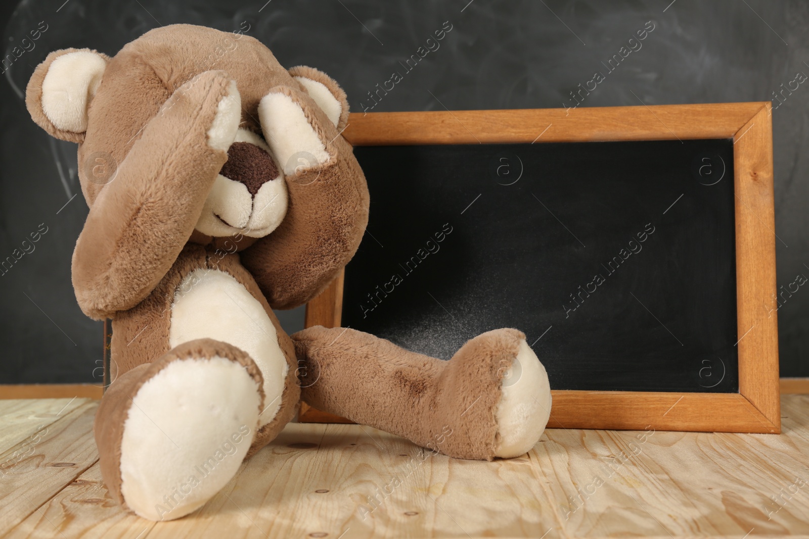 Photo of Teddy bear covering eyes and small blackboard on wooden table. Space for text