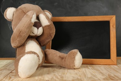 Photo of Teddy bear covering eyes and small blackboard on wooden table. Space for text