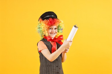 Happy little boy dressed like clown with confetti popper on yellow background. Surprise party