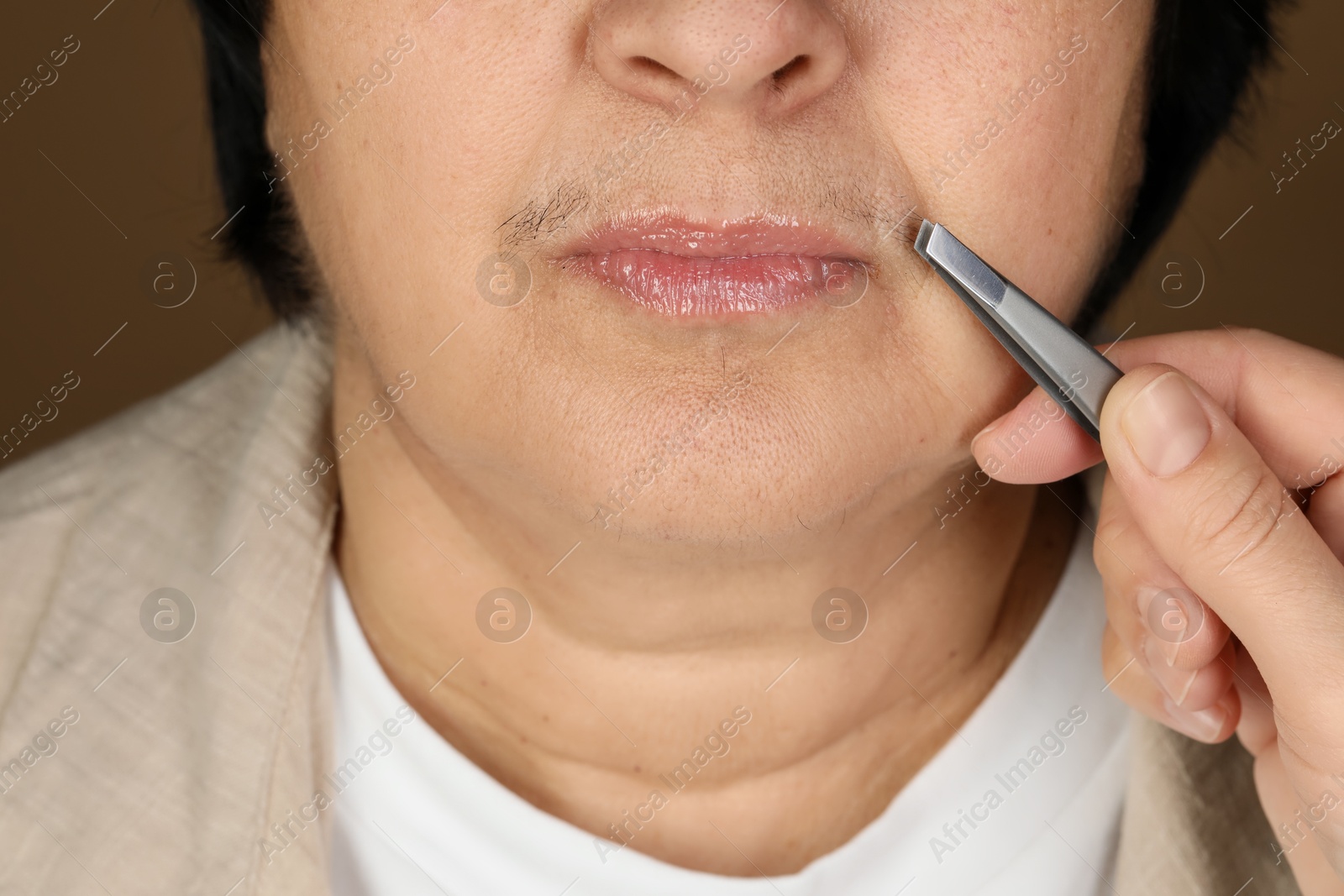 Photo of Senior woman plucking her mustache with tweezers on brown background, closeup