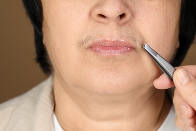 Photo of Senior woman plucking her mustache with tweezers on brown background, closeup