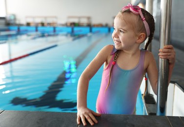 Little girl with goggles getting out of swimming pool indoors, space for text