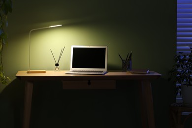 Photo of Comfortable workplace with laptop and lamp on wooden desk at home office in evening