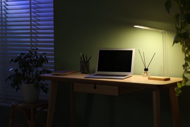 Photo of Comfortable workplace with laptop and lamp on wooden desk at home office in evening