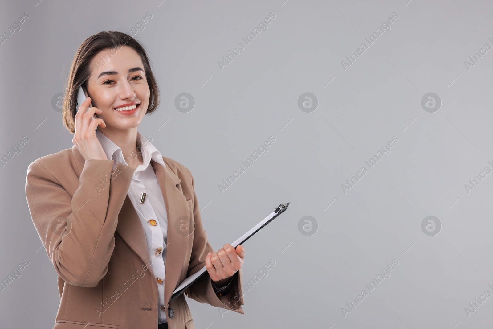 Photo of Smiling secretary with clipboard talking on smartphone against grey background. Space for text