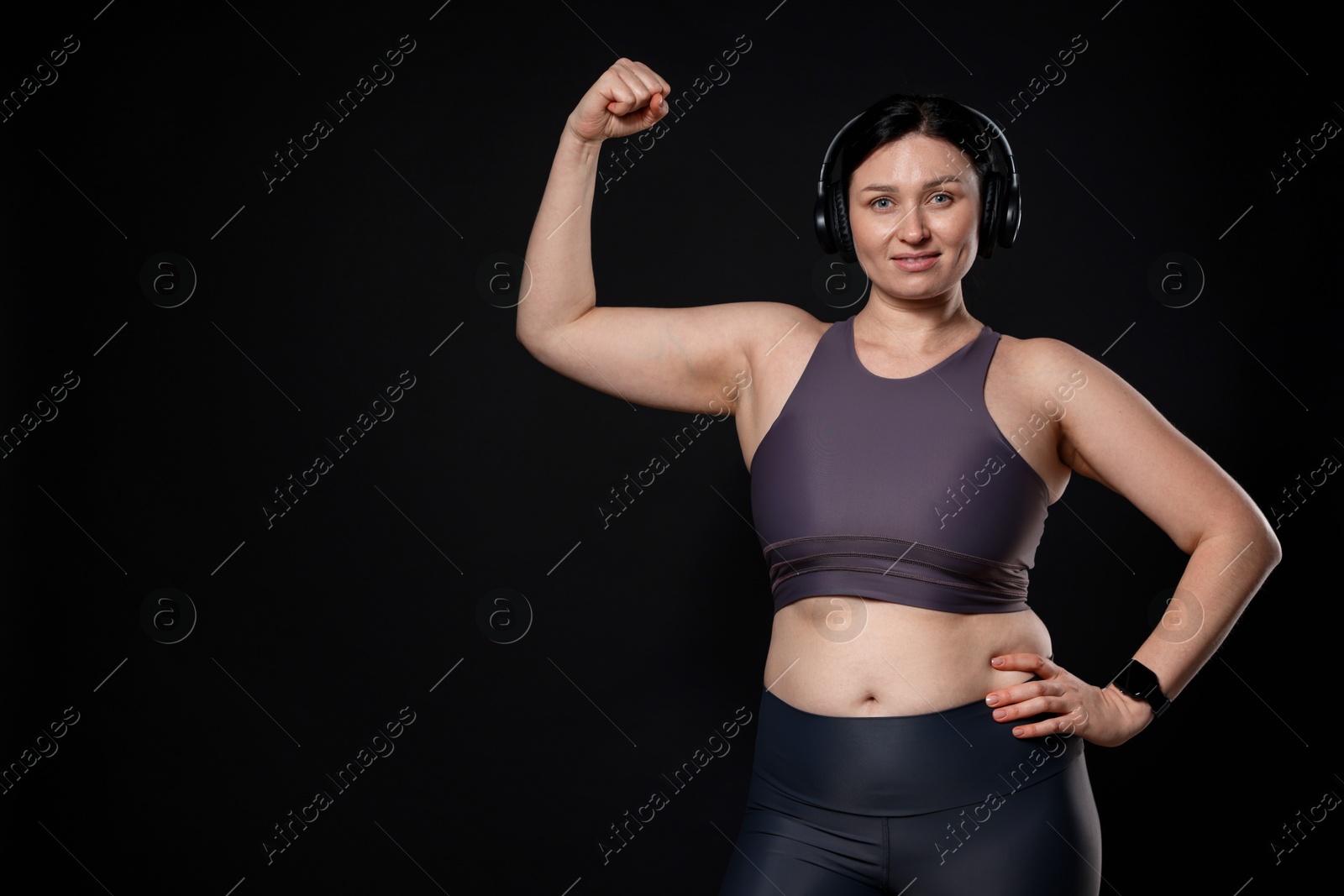 Photo of Plus size woman in gym clothes and headphones showing her bicep on black background, space for text