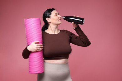 Plus size woman in gym clothes with fitness mat and water bottle on pink background