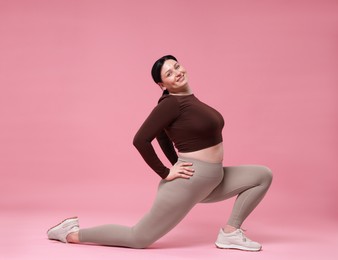 Plus size woman in gym clothes exercising on pink background