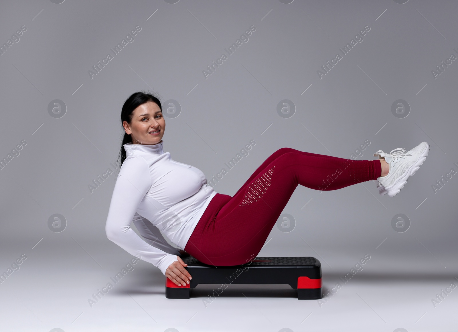 Photo of Plus size woman in gym clothes doing exercise with step platform on grey background