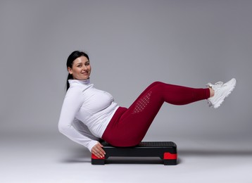 Plus size woman in gym clothes doing exercise with step platform on grey background