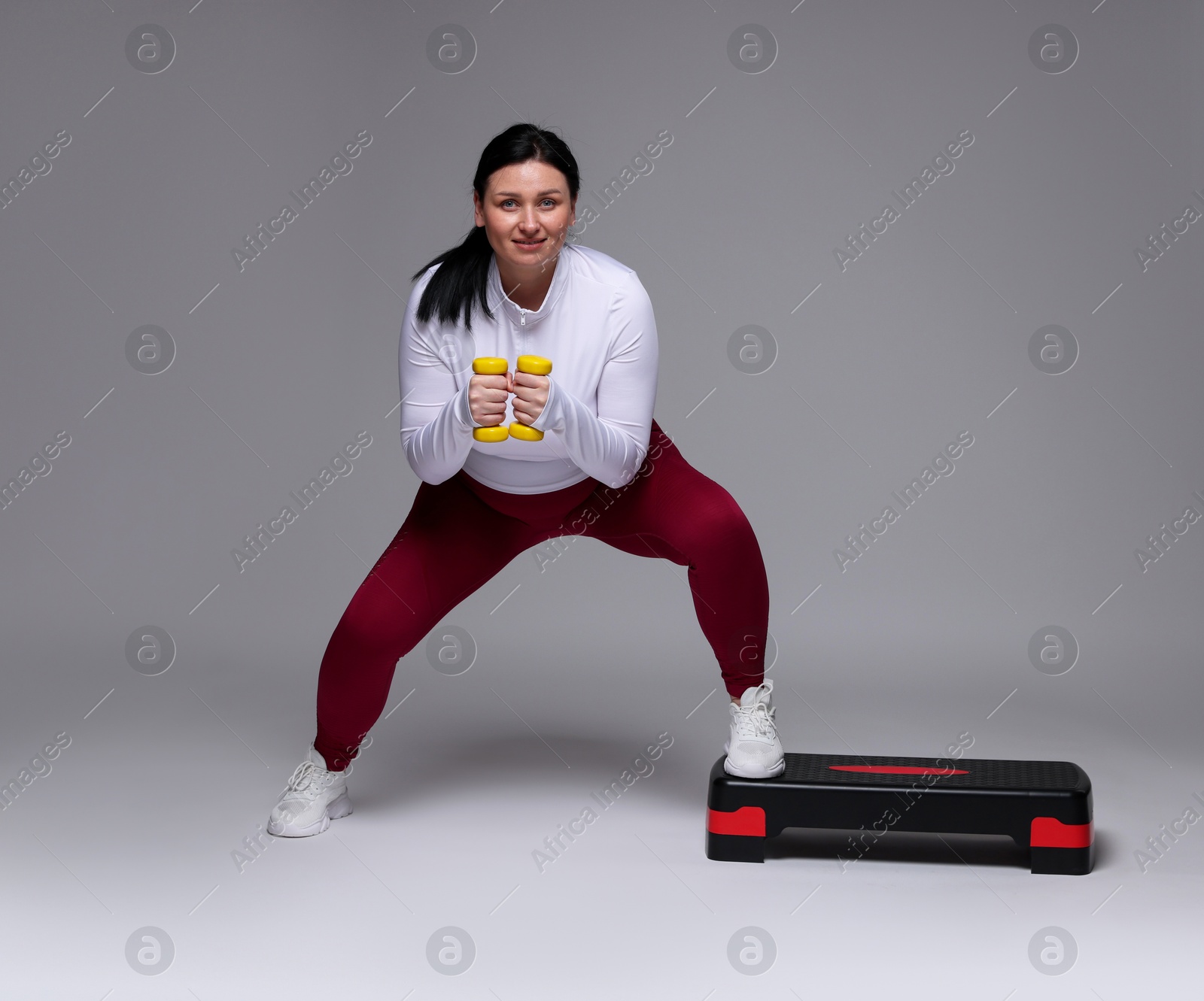 Photo of Plus size woman in gym clothes doing exercise with dumbbells and step platform on grey background