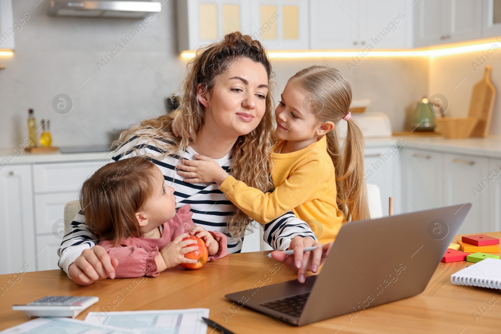 Photo of Single mother working on laptop and her daughters at table in kitchen