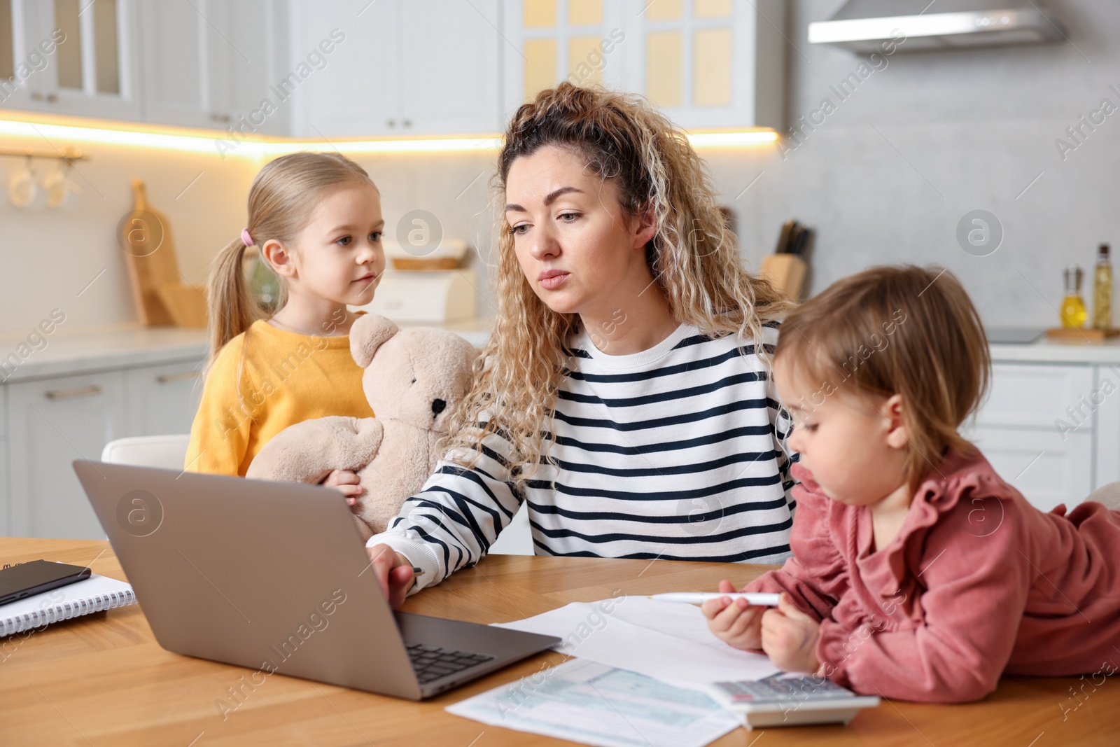 Photo of Single mother working on laptop and her daughters at table in kitchen