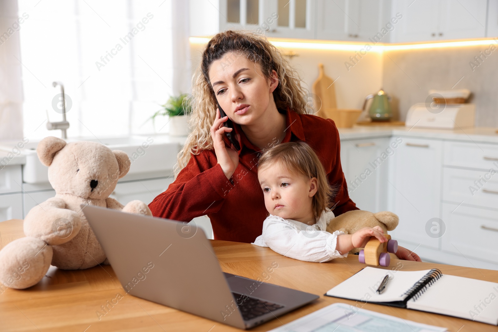 Photo of Work-family balance. Single mother holding her daughter while talking on smartphone at table in kitchen