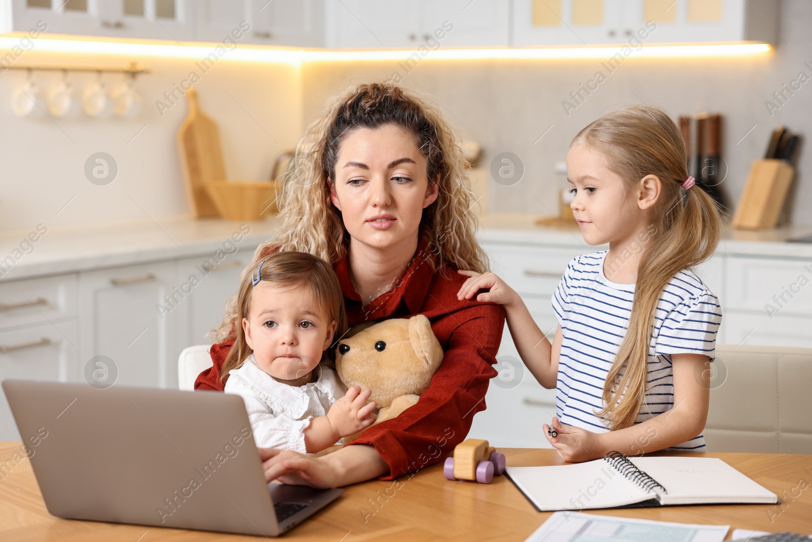 Photo of Single mother working with laptop and her daughters at table in kitchen