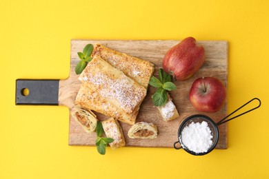 Photo of Tasty apple strudels with powdered sugar, fruits and mint on yellow background, top view