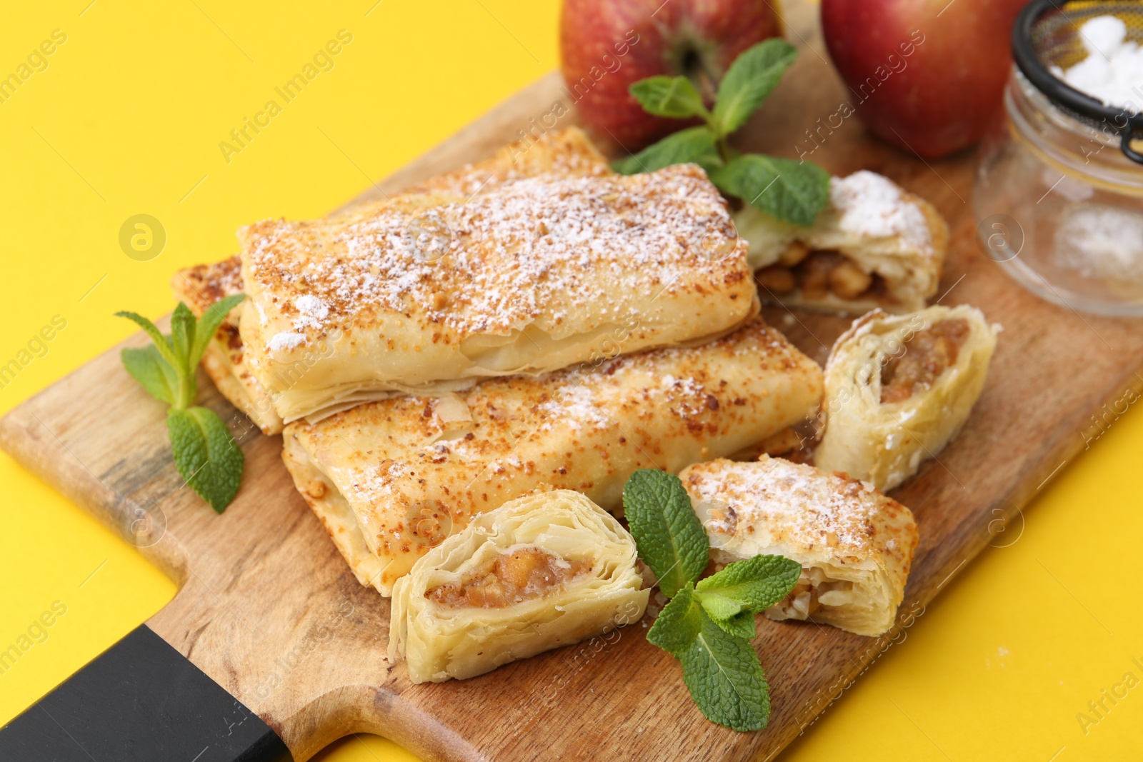 Photo of Tasty apple strudels with powdered sugar, fruits and mint on yellow background, closeup