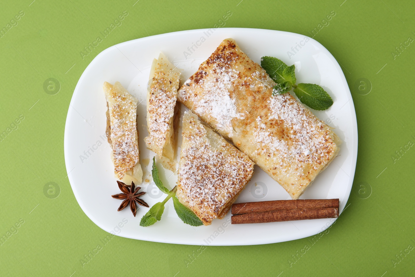 Photo of Tasty apple strudels with powdered sugar, mint and spices on green background, top view