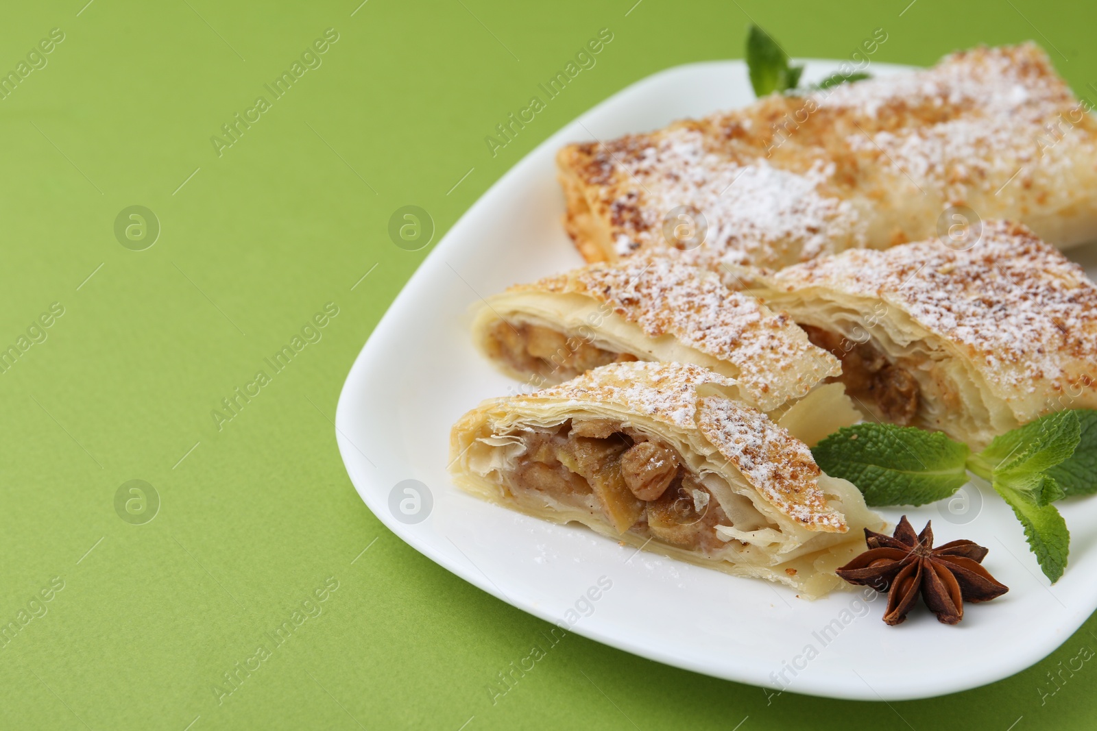 Photo of Tasty apple strudels with powdered sugar, mint and anise on green background, closeup. Space for text