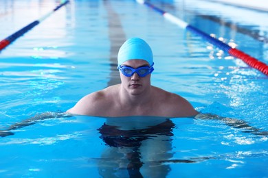 Young man in cap, goggles and swimwear swimming in pool indoors