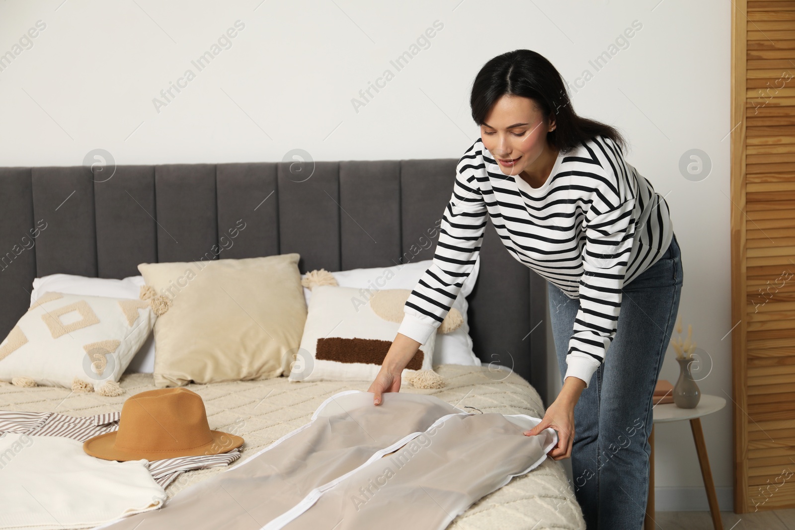 Photo of Woman taking garment cover with clothes from bed indoors