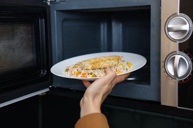 Photo of Woman putting plate with lunch into microwave indoors, closeup