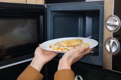 Photo of Woman putting plate with lunch into microwave indoors, closeup