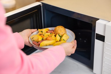 Photo of Woman putting plate with lunch into microwave indoors, closeup