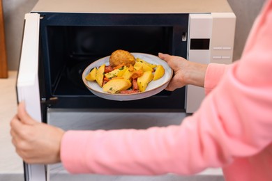 Photo of Woman putting plate with lunch into microwave indoors, closeup